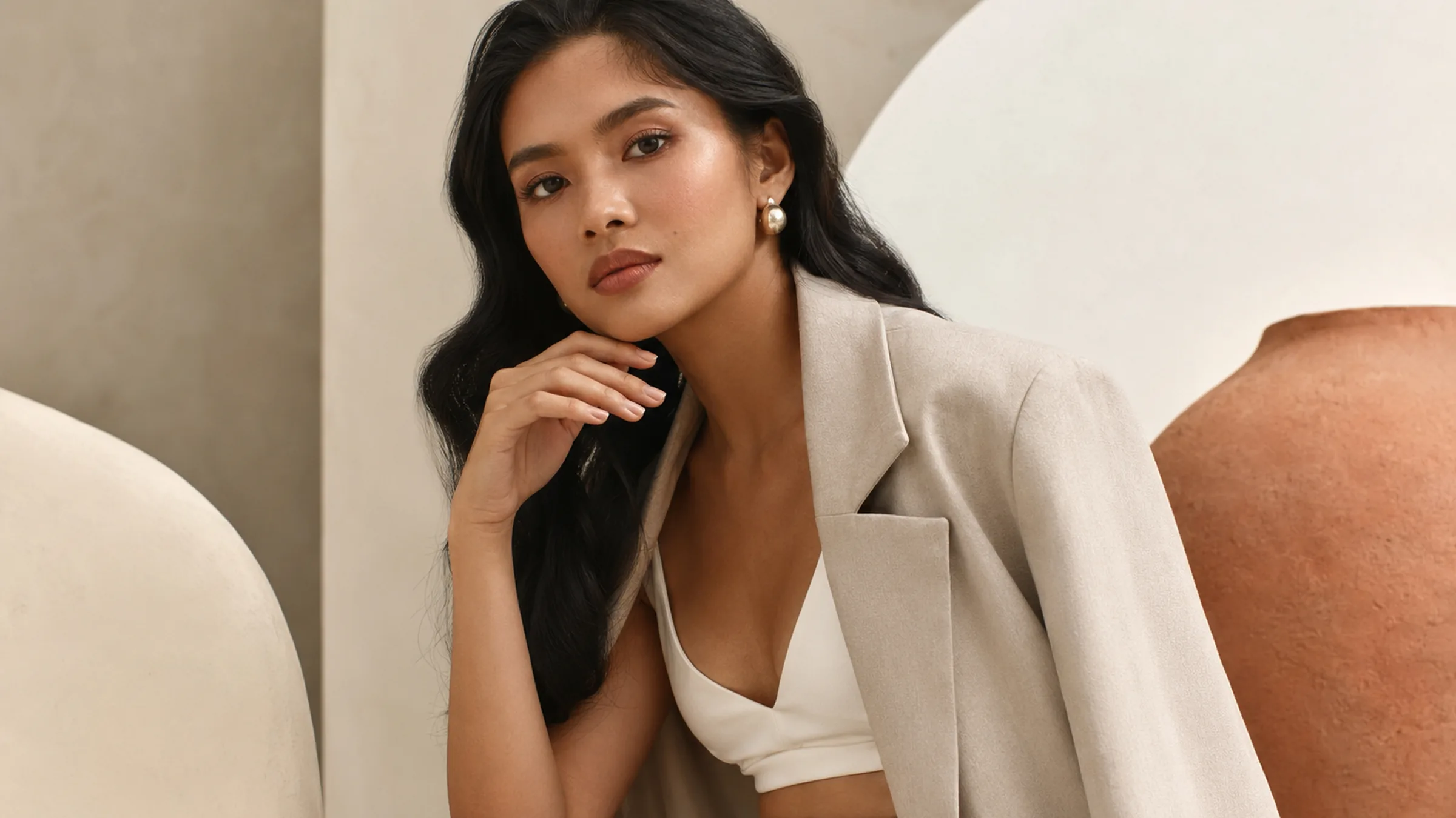 A woman in a stone blazer, her hand resting at her chin, wearing a pearl earring against a warm beige wall.